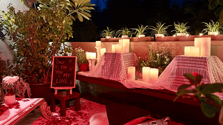 An intimate outdoor seating setup with floor cushions, a low table, flower petals, candles, potted plants, and a 'Only for you my love' sign.