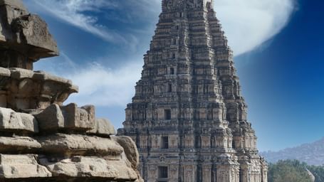 A wide-angle, low-angle shot of a tall stone temple (gopuram) with multiple smaller towers, surrounded by other temple structures and a rocky, arid landscape under a dramatic blue sky with wispy clouds.