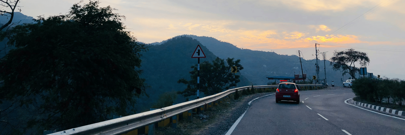 Scenic mountain road in Kasauli at sunset, with a car driving along a winding highway surrounded by rolling hills and a colourful evening sky.