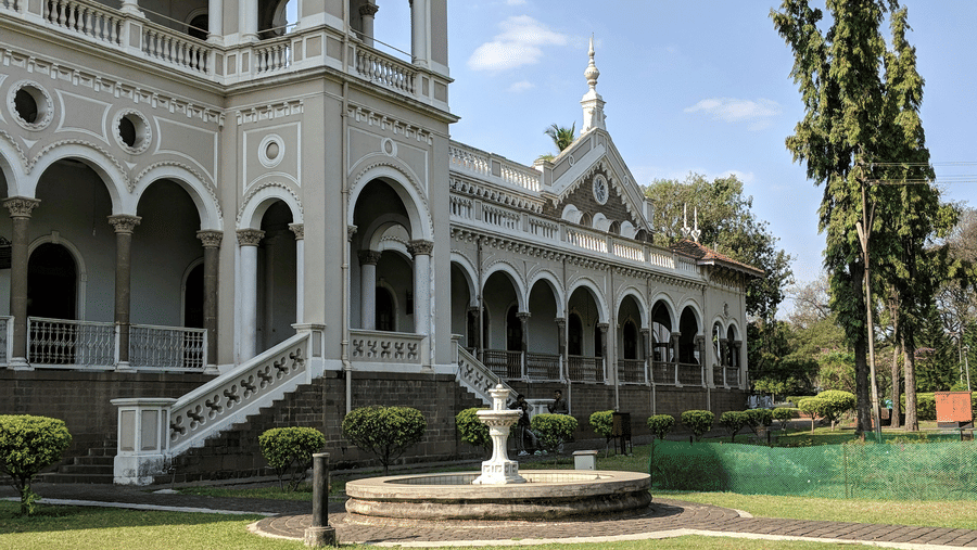 A facade view of Aga Khan Palace showcases arched corridors, upper balconies and an open lawn with a sculpted fountain.