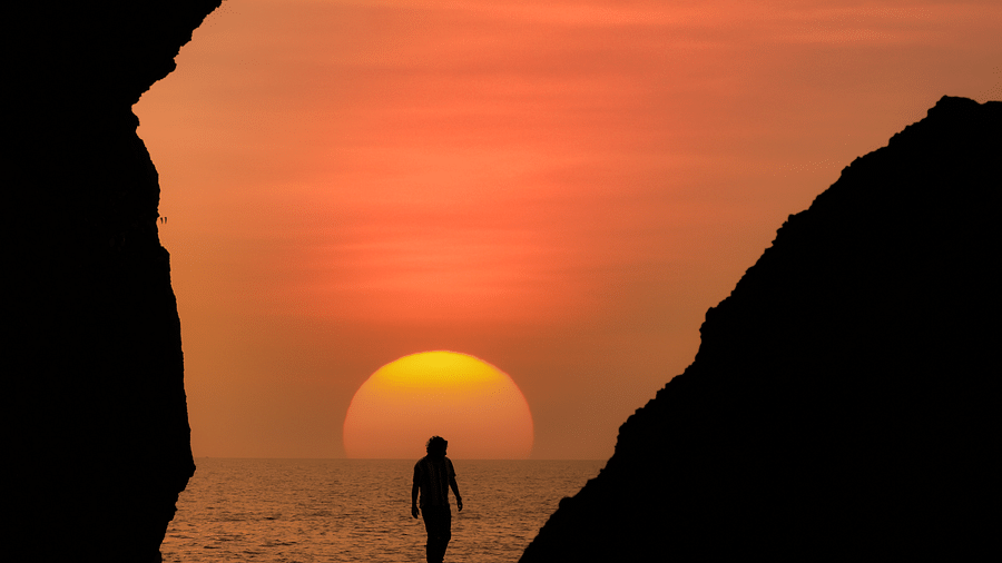A silhouette of a person standing on a rock or cliff overlooking a body of water, with a large, bright orange sunset filling the sky behind them, creating a dramatic, warm glow.