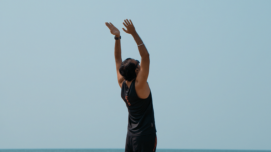 A person in dark clothing stands on a sandy beach facing the ocean, with arms raised above their head in a yoga pose, possibly mountain pose or a stretch.
