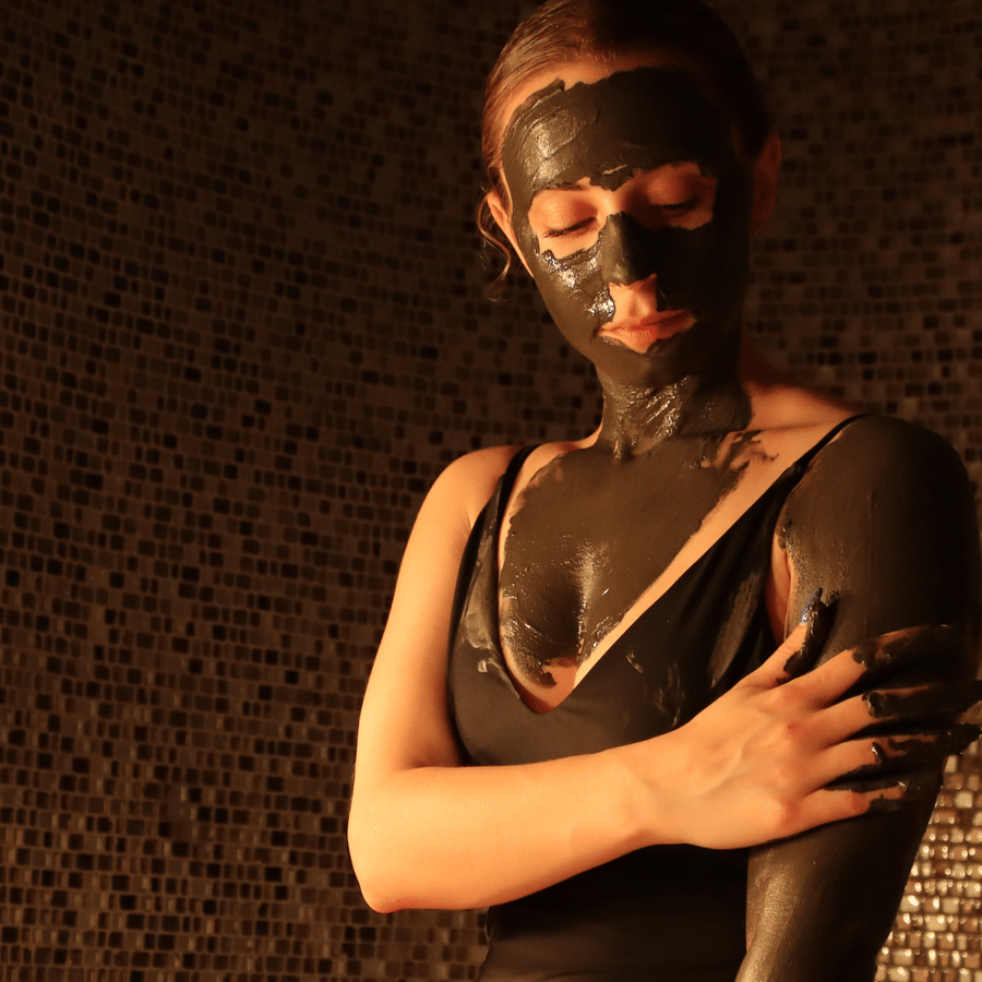 A woman with a mud mask on her face and mud applied to her arm, in a dark, earthy-toned spa setting at YO1 Longevity & Health Resorts