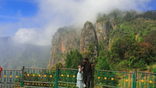 Couple standing by a railing overlooking mist-covered cliffs and lush green hills in Kodaikanal, with dramatic rock formations rising through the clouds.