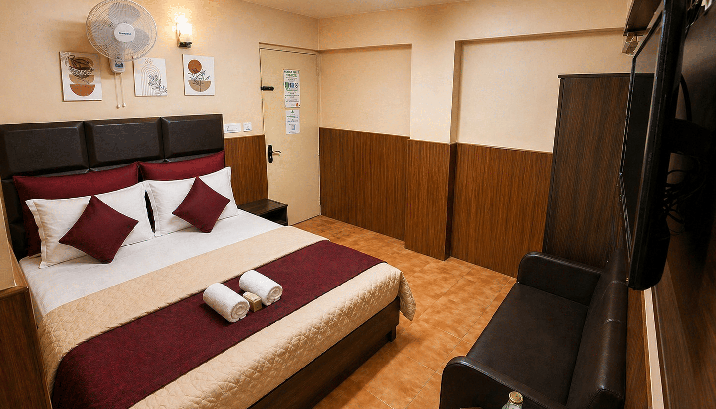 A neatly-made bed, a leather sofa, and a wall-mounted television across from the bed in the Deluxe Double Room With Extra Cot at BN Tourist Home, Ooty.