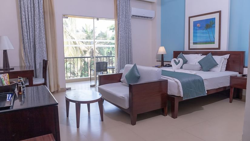 Complete view of the Deluxe room with a double bed, a couch a small tea table and a balcony at Kyriad Prestige Calangute, Goa