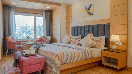 A bedroom with a king size bed, patterned bedding, bedside tables, a bench, 2 chairs, a lamps, wooden flooring, and a window at Perfectstayz Premium At Mall Road (Hotel Super), Mussoorie.