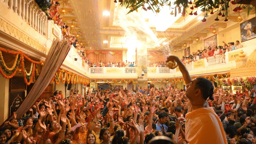 A brightly lit religious gathering inside a temple with a large crowd and a person holding up an arm.