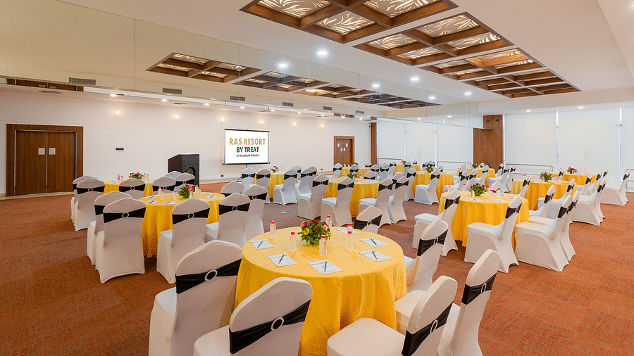 A side angle view of a spacious banquet hall at Ras Resort by Treat, Silvassa, with round tables covered in tablecloths and white chair covers, ready for a corporate event or conference.