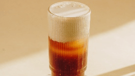 tall glass of a layered iced coffee drink with a clear bottom layer and a white top layer, with a few coffee beans nearby at Gemini Continental