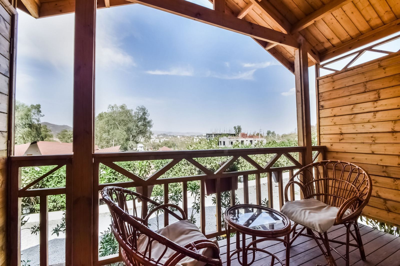A wooden balcony with two wicker chairs and a small table overlooks a scenic view of trees .- Bamboo Saa Resort & Spa, Udaipur