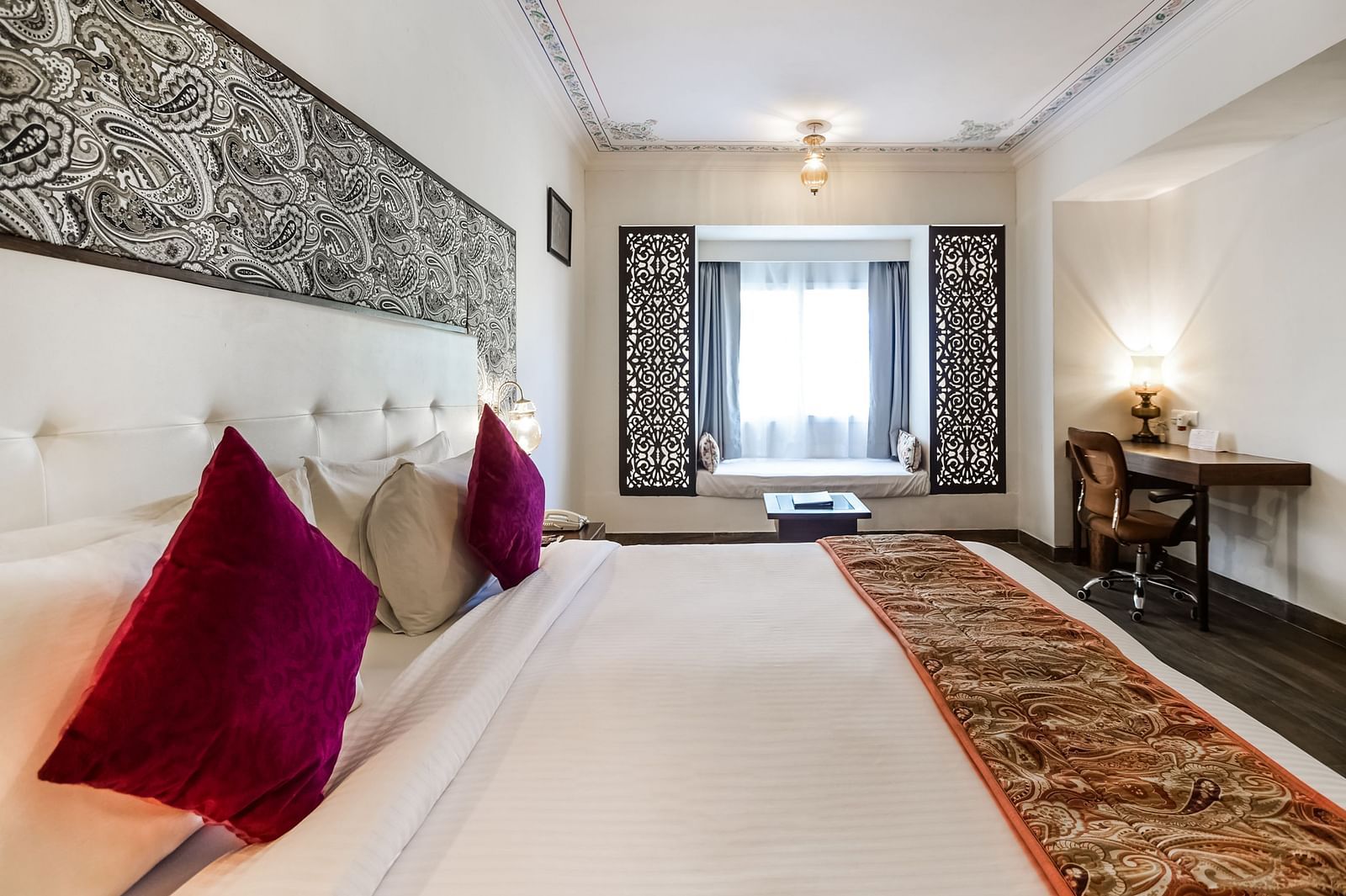 A hotel room with a plush bed, decorative wall panel, a window seat, and a desk.- Bamboo Saa Resort & Spa, Udaipur