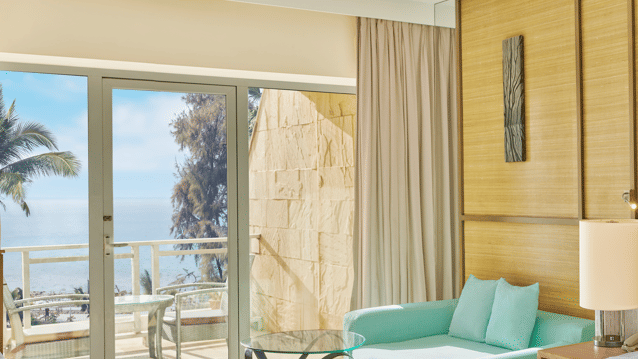 Bright, airy room with a mint-green sofa, glass coffee table, and a balcony offering a clear view of the sea and palm trees at The Resort, Mumbai, the perfect place for a Couple Staycation near Mumbai