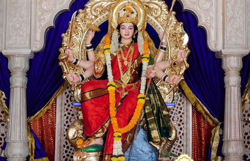 Idol of a goddess dressed in red and green attire, adorned with a golden crown, multiple arms holding weapons, seated on a decorated throne with garlands.
