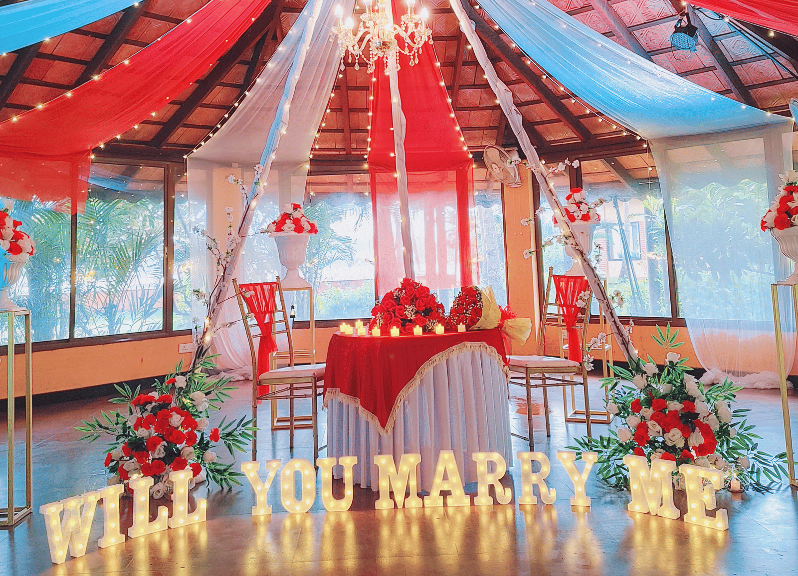 'Will You Marry Me' proposal decoration in an indoor set up with red and white drapes and floral arrangements at Paradise Lagoon Resort, Udupi.