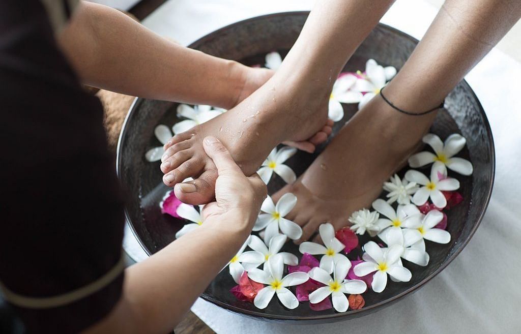 A spa therapist is gently massaging a client's feet, which are soaking in a bowl of water filled with white flower petals.