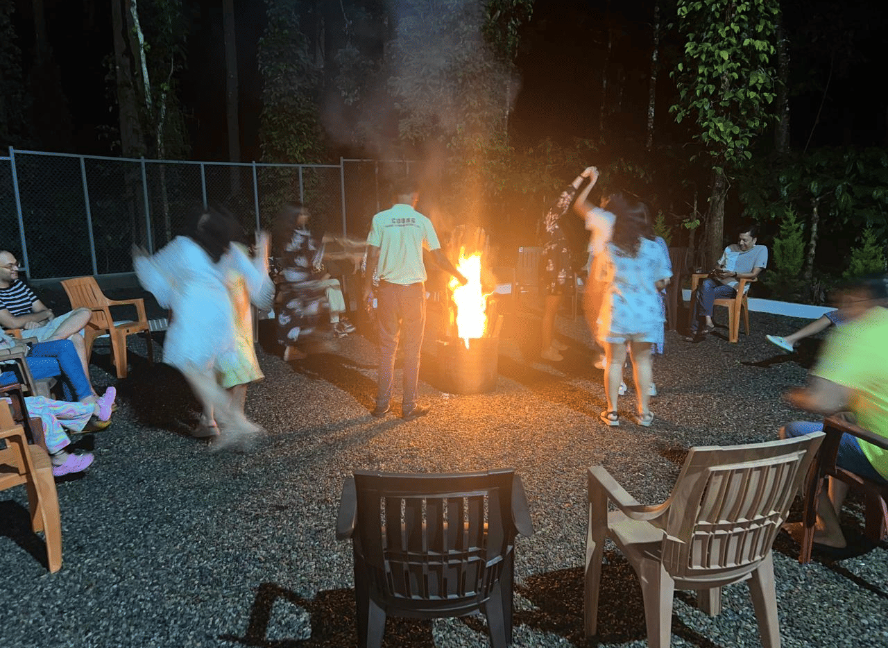 A group of people socialising and dancing around a large bonfire at night, with several plastic chairs surrounding the fire pit at Coorg Orange Blossom Resort & Spa.