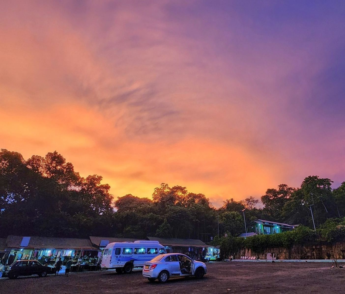 A fiery orange and purple sky at sunset over a car park with trees and a few cars.