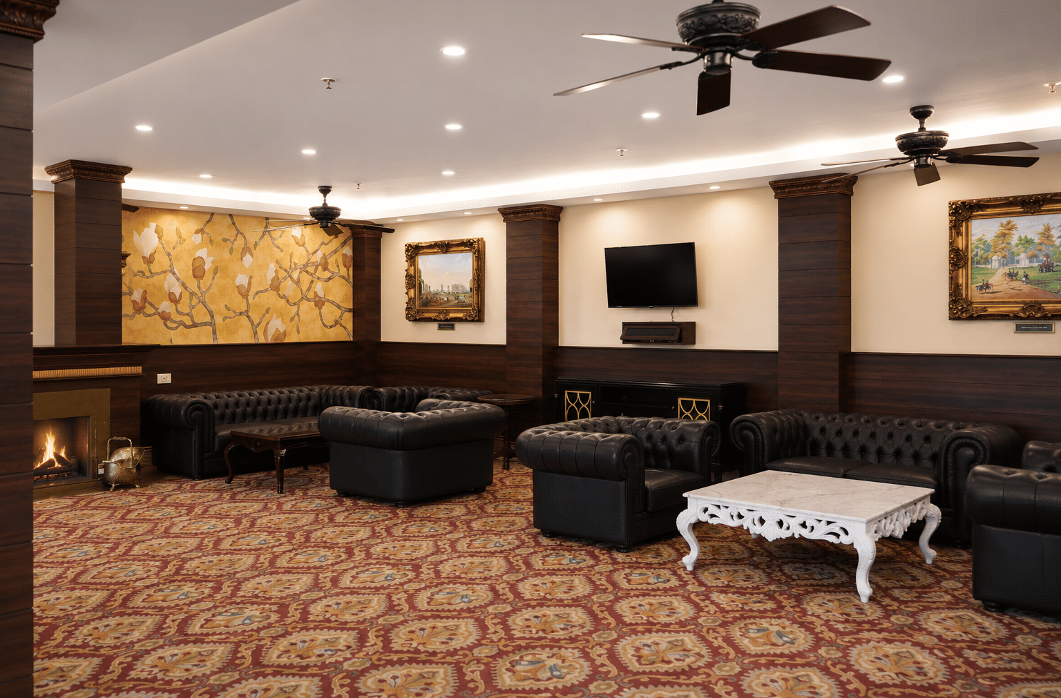 Drawing Room lounge with black leather Chesterfield sofas, a lit fireplace, a floral wall mural and a carved coffee table at MAYFAIR Himalayan Spa Resort, Kalimpong.