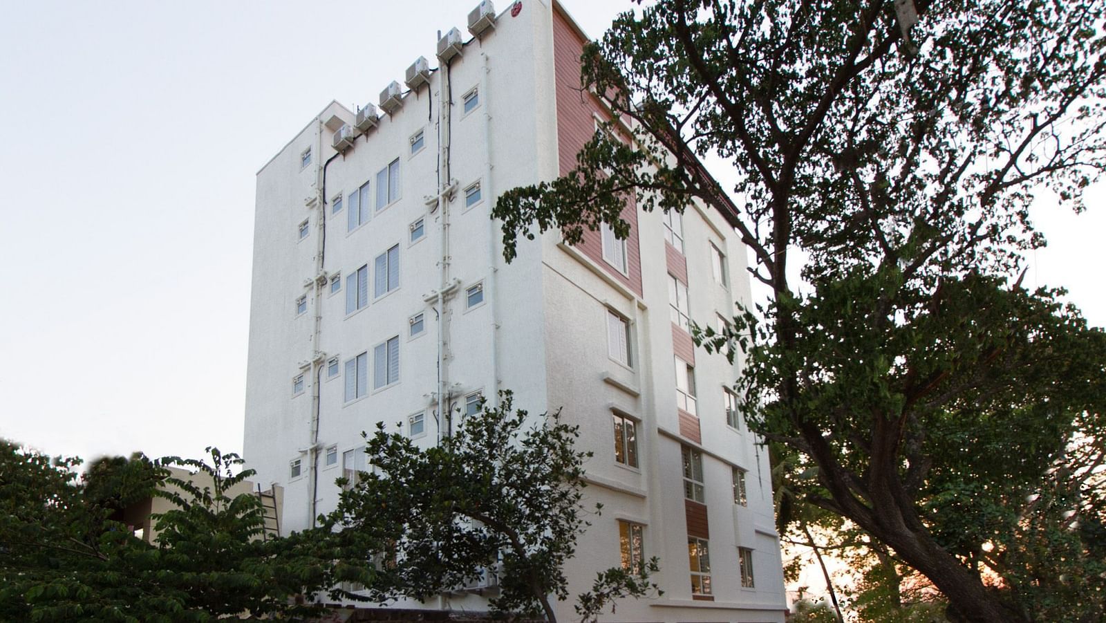 The multi-storey building of Sanctum Suites Whitefield, Bangalore amongst trees under a daytime sky.