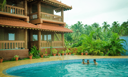 A multi-storey wooden building overlooking a turquoise swimming pool with guests enjoying the water at Paradise Lagoon Resort, Udupi.