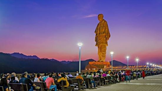 Tourists gather at the base of the Statue of Unity, surrounded by evening lights and scenic hills under a colourful twilight sky.