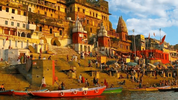 Crowds of people and colourful boats gather along the iconic stone steps of Assi Ghat beside old historic buildings in Varanasi.