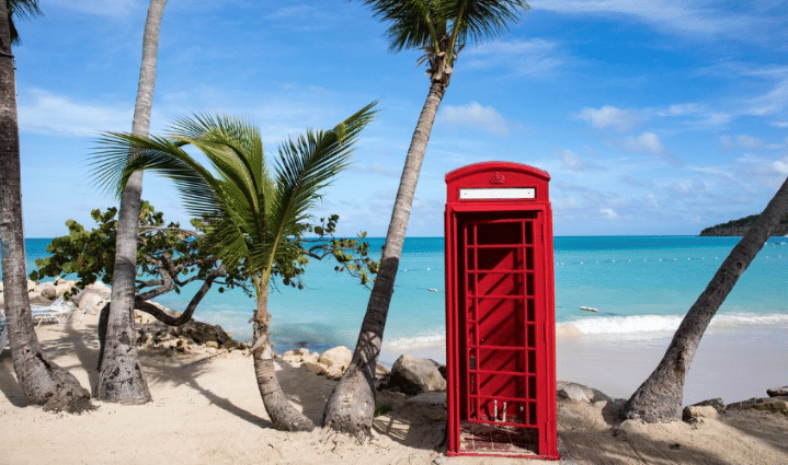 A vibrant red British telephone box stands on a white sand beach lined with palm trees, overlooking the turquoise ocean under a clear blue sky.