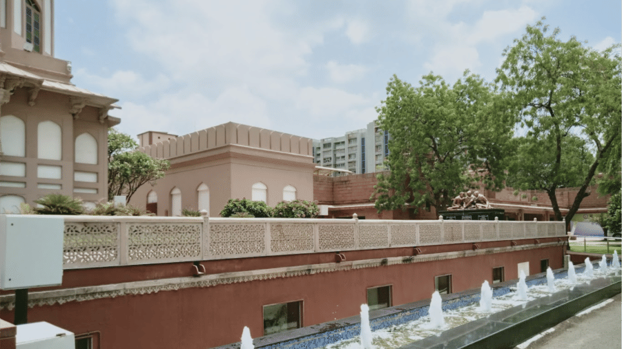 The exterior view of Sardar Vallabhbhai Patel National Memorial featuring arched windows, elevated walkway, trees, and a series of small water fountains along its base.