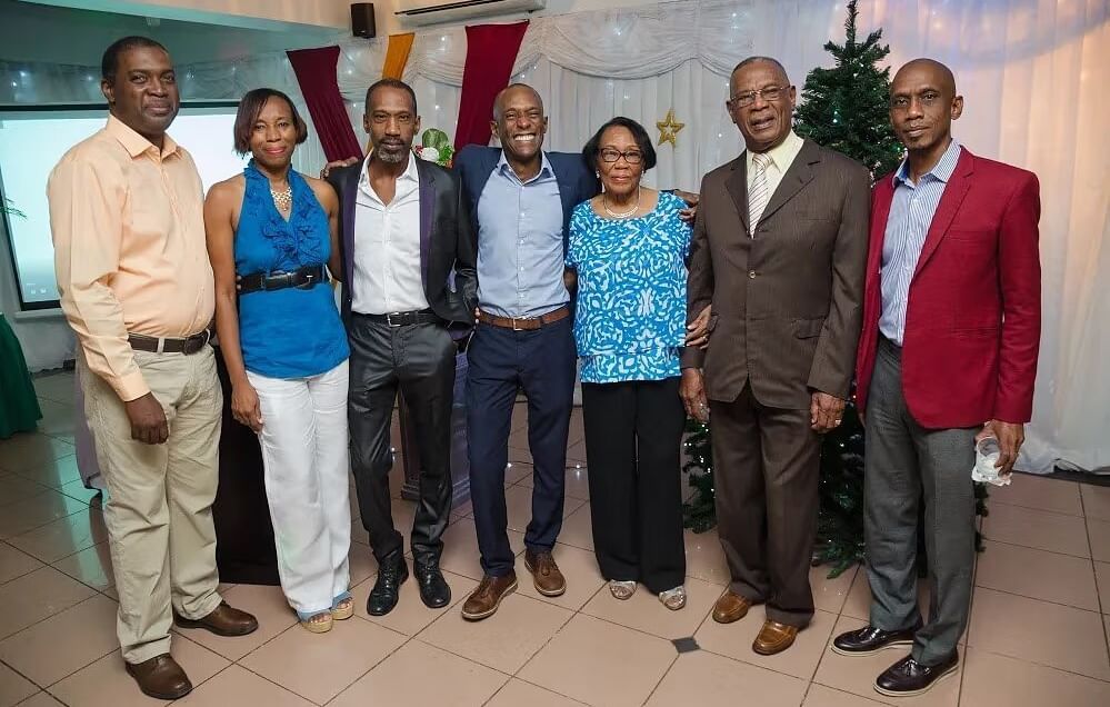 A group of seven formally dressed individuals, five men and two women, stand together smiling in an indoor setting.