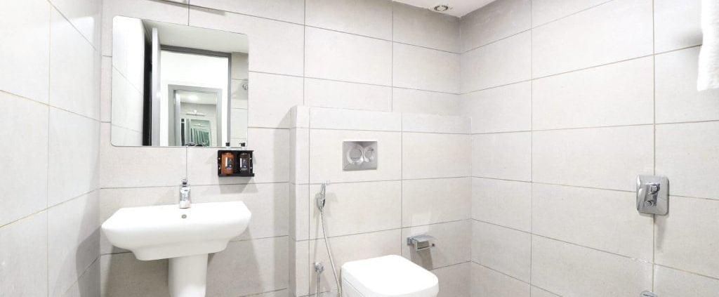 A bathroom with white tiles all around featuring wash basin along with a commode and a shower area at Pravasa Ahmedabad.