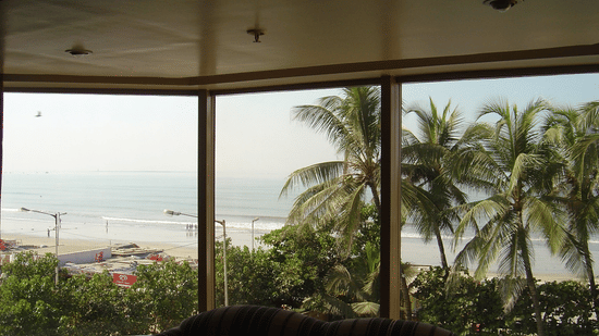 Scenic view of a beach and the sea through a big hotel room window, framed by lush palm trees.