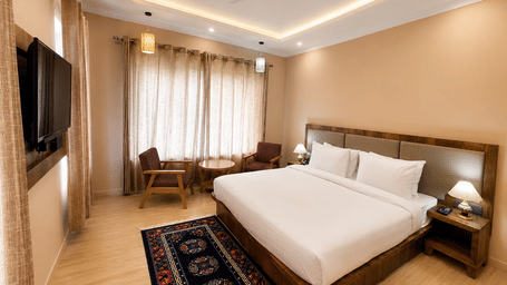 Hotel room with a bed dressed in white linen, a wooden headboard, a wall-mounted television, a patterned rug and warm ambient lighting nearby.