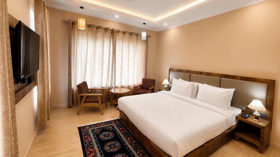 Hotel room with a bed dressed in white linen, a wooden headboard, a wall-mounted television, a patterned rug and warm ambient lighting nearby.