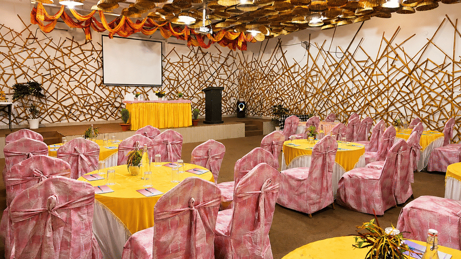 anquet hall with round tables dressed in yellow linen, pink patterned chair covers, a decorative bamboo lattice ceiling and a stage area nearby.