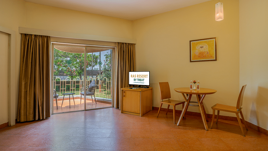 A spacious living room at Ras Resort by Treat, Silvassa, featuring a seating area with a table and chairs, a television, and a balcony overlooking the garden.