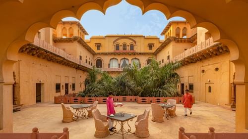 The central courtyard of Neemrana's Tijara Fort Palace, viewed through an ornate archway, showcases seating areas and lush greenery within the historic structure.