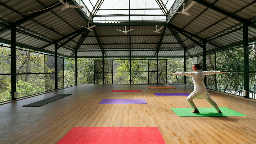 A person practising yoga on a colourful mat in a spacious, glass-walled pavilion surrounded by lush greenery and trees at Neemrana's Glasshouse on The Ganges.