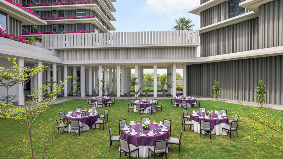 Lawn set with round tables in purple and white linen and grey chairs, enclosed by a colonnade and flowering tower balconies at The Residency Signature Madurai.