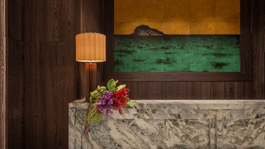 Reception desk in green-veined marble with a floral arrangement, a pleated lamp and a framed sea painting on a wooden wall at The Residency Signature Madurai.
