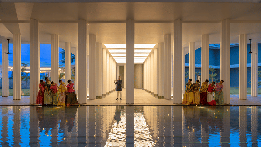 2 groups of women in traditional skirts by the pillars of a lit colonnade, a flautist and candles on the reflecting pool at The Residency Signature Madurai.