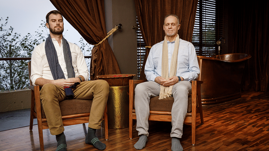 Two men sit side-by-side in wooden armchairs indoors, meditating with their eyes closed.