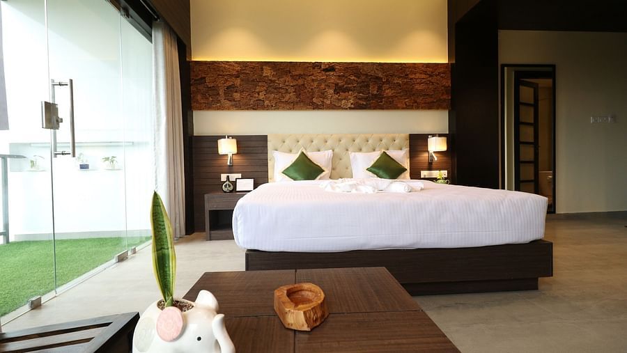 A hotel bedroom with a large bed, a small table in the foreground, and a view of an outdoor green space through a large glass door - Sea Hills Hotels, Sri Vijaya Puram (Port Blair)