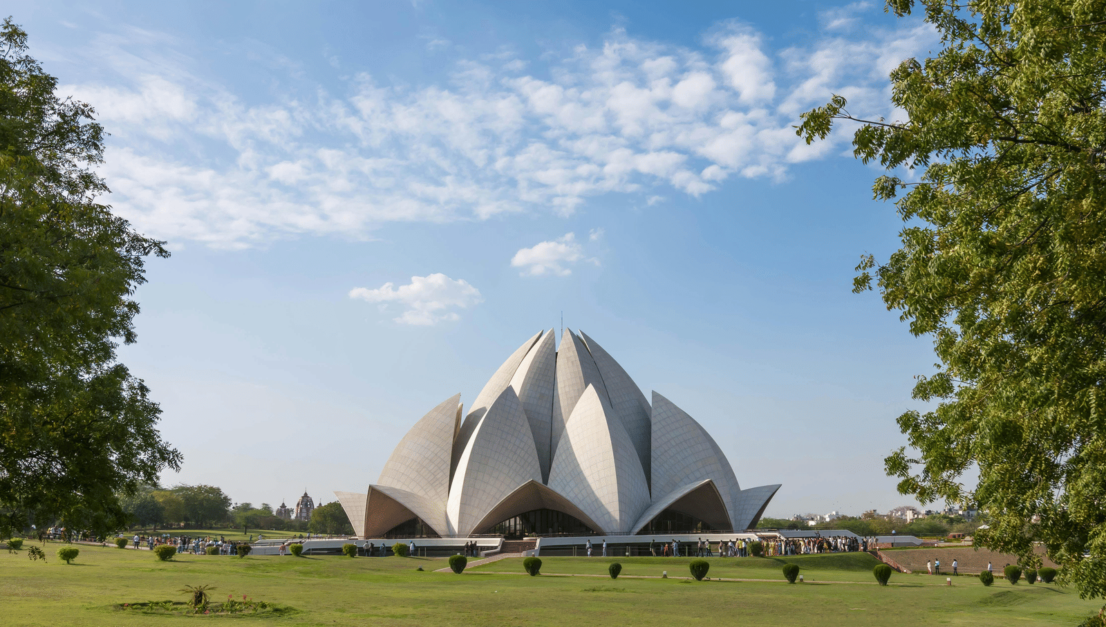 Facade shot of Lotus Temple surrounded by lush greenery and backdrop of clear blue sky.