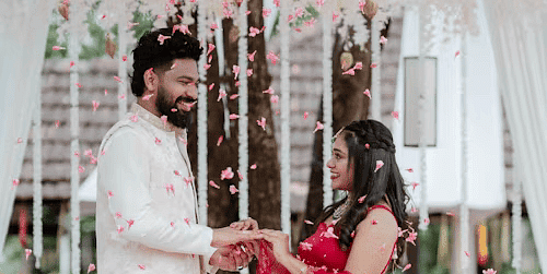 Romantic proposal moment under floral decor, with groom kneeling before bride in red bridal attire.