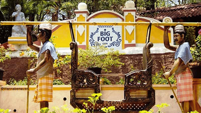 The entrance of Big Foot museum with a yellow sign, flanked by two statues wearing traditional clothes and hoisting a palanquin.