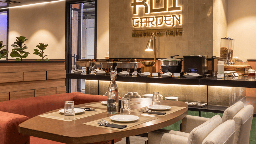 A close-up view of a dining layout inside Koi Garden restaurant. A table is set with plates, cutlery, and glasses between chairs and a booth. The restaurant logo is visible on the background wall.