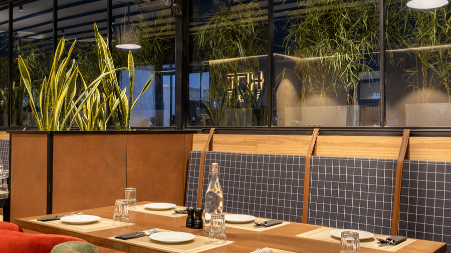 A close-up view of a set dining table at Koi Garden inside Hotel Bawa Gurgaon. Plates and cutlery are arranged on the surface with chairs positioned around it. A hanging lamp illuminates the area from above.