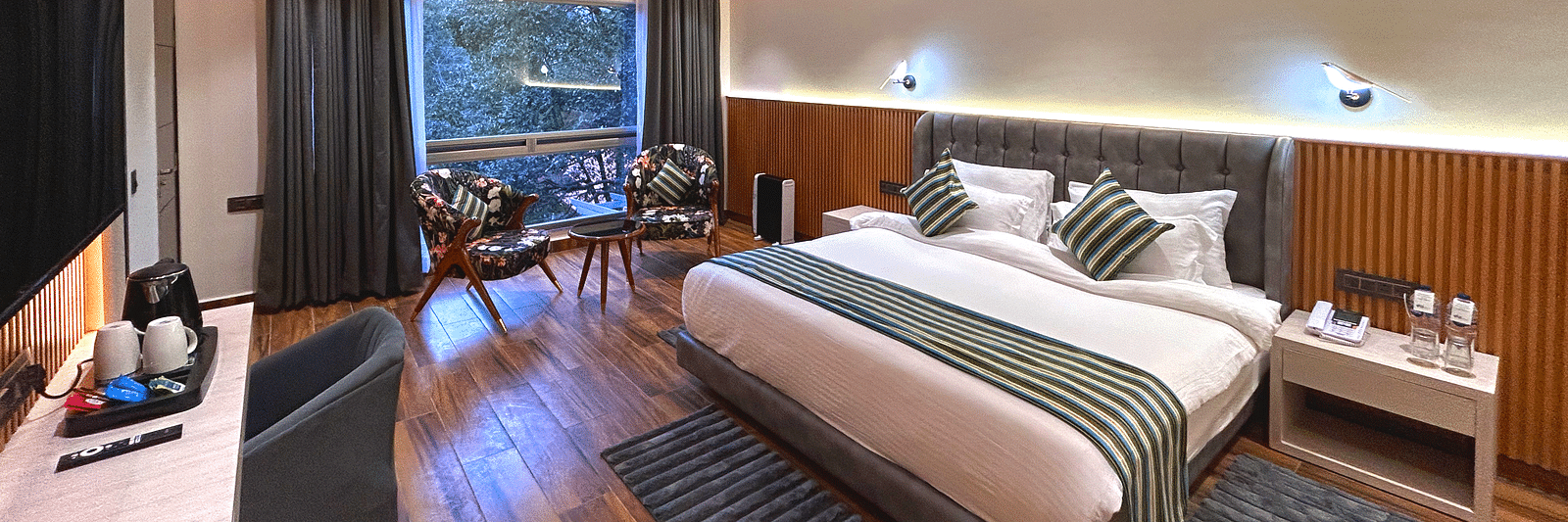The premium room of DLS The Emerald, Nainital with a bed, bed side table, chairs, coffee table, a window overlooking the view, with warm lighting
