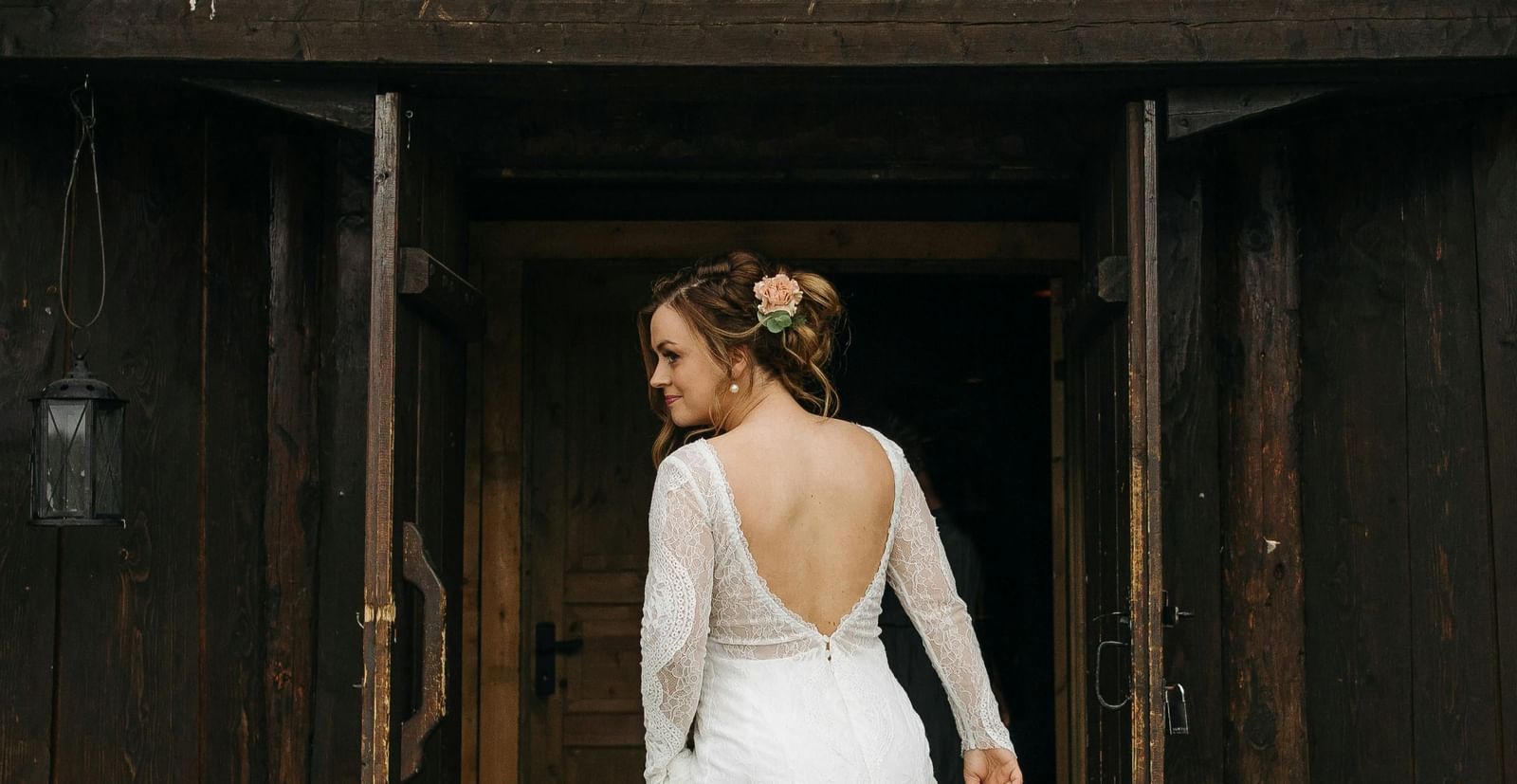 A bride in a long-sleeved, open-back gown stands by a rustic building, a beautiful scene near the Tallman Hotel.